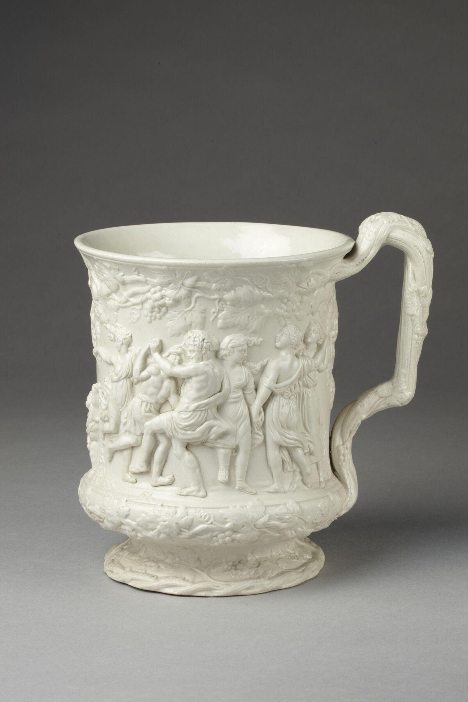 White relief-molded ceramic mug depicting a crowded bacchanalian scene with satyrs, maenads, and putti in ecstatic movement, with a curved handle.