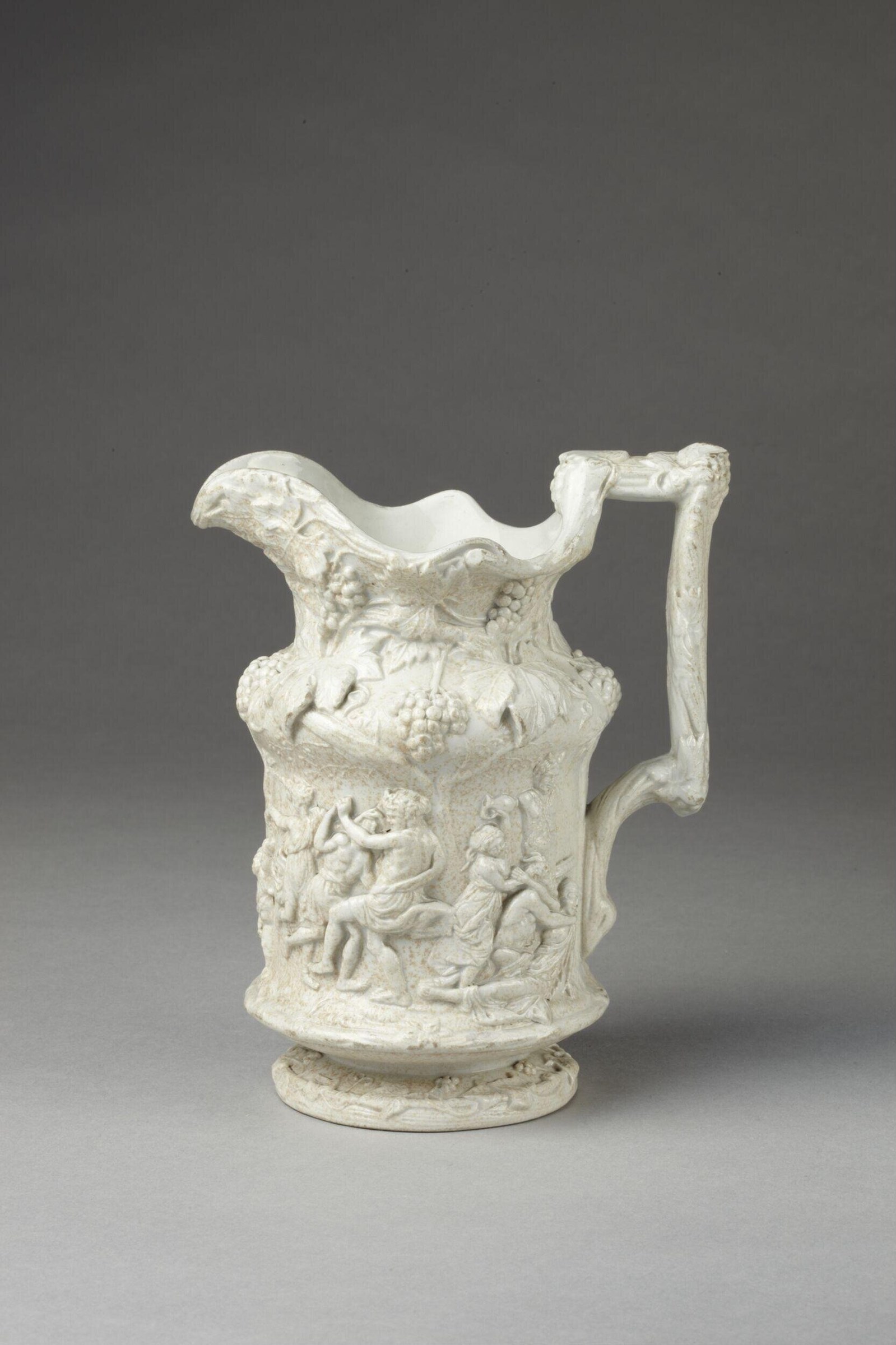 White ceramic jug with dense relief carvings of intoxicated figures, satyrs, and grapevines covering its body.