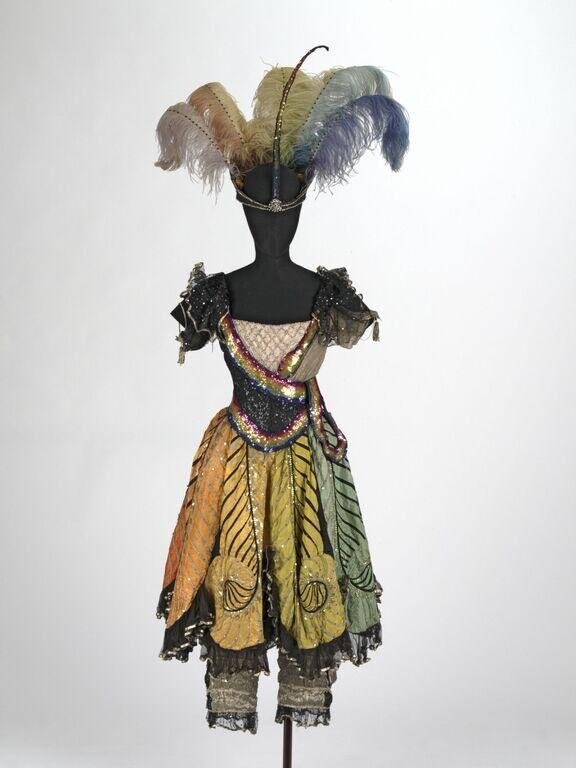 Black bodice with jeweled sash and multicolored feathered silk skirt with lace trim, topped by arched ostrich plumes in cream, gold, green, and blue.