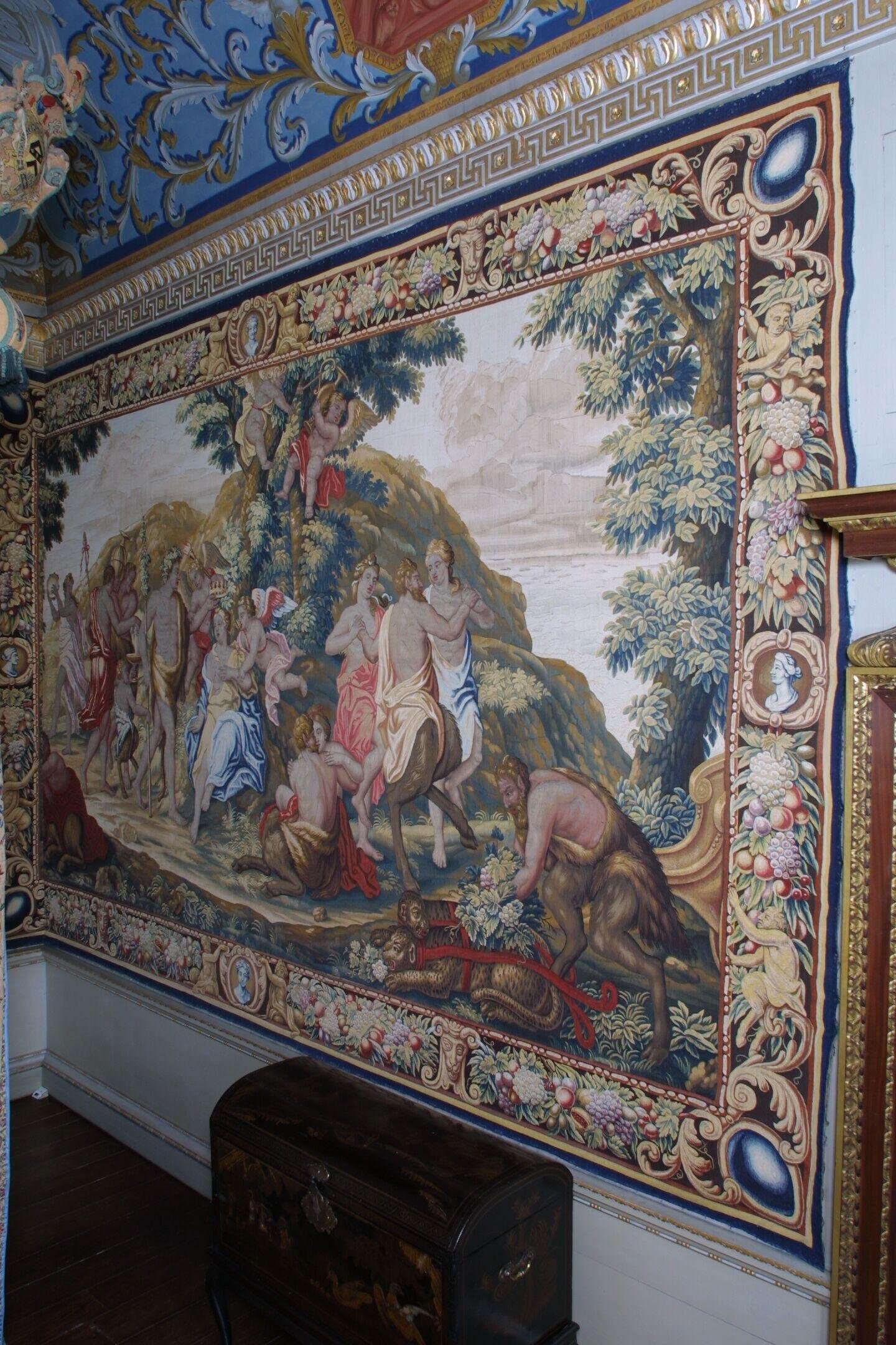 Baroque tapestry depicting a mythological scene with an elaborately dressed group of figures gesturing from an elevated rocky formation at left, while a