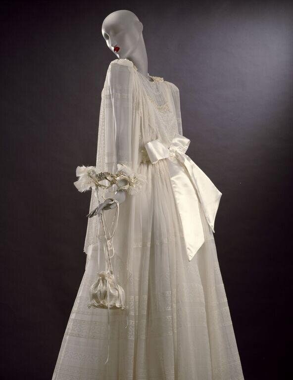 Cream silk evening dress with layered pleats and back bow, displayed on a mannequin against dark background.