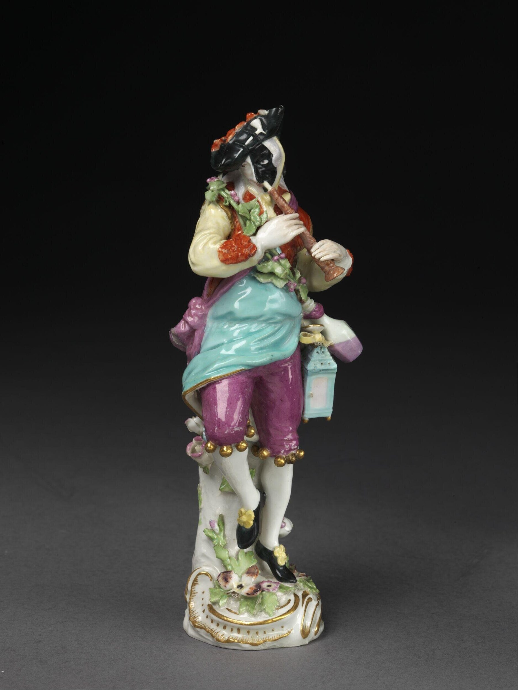 Porcelain figurine of a masked dancer in turquoise and magenta silk costume, holding strawberries and a lantern, on a rococo base.