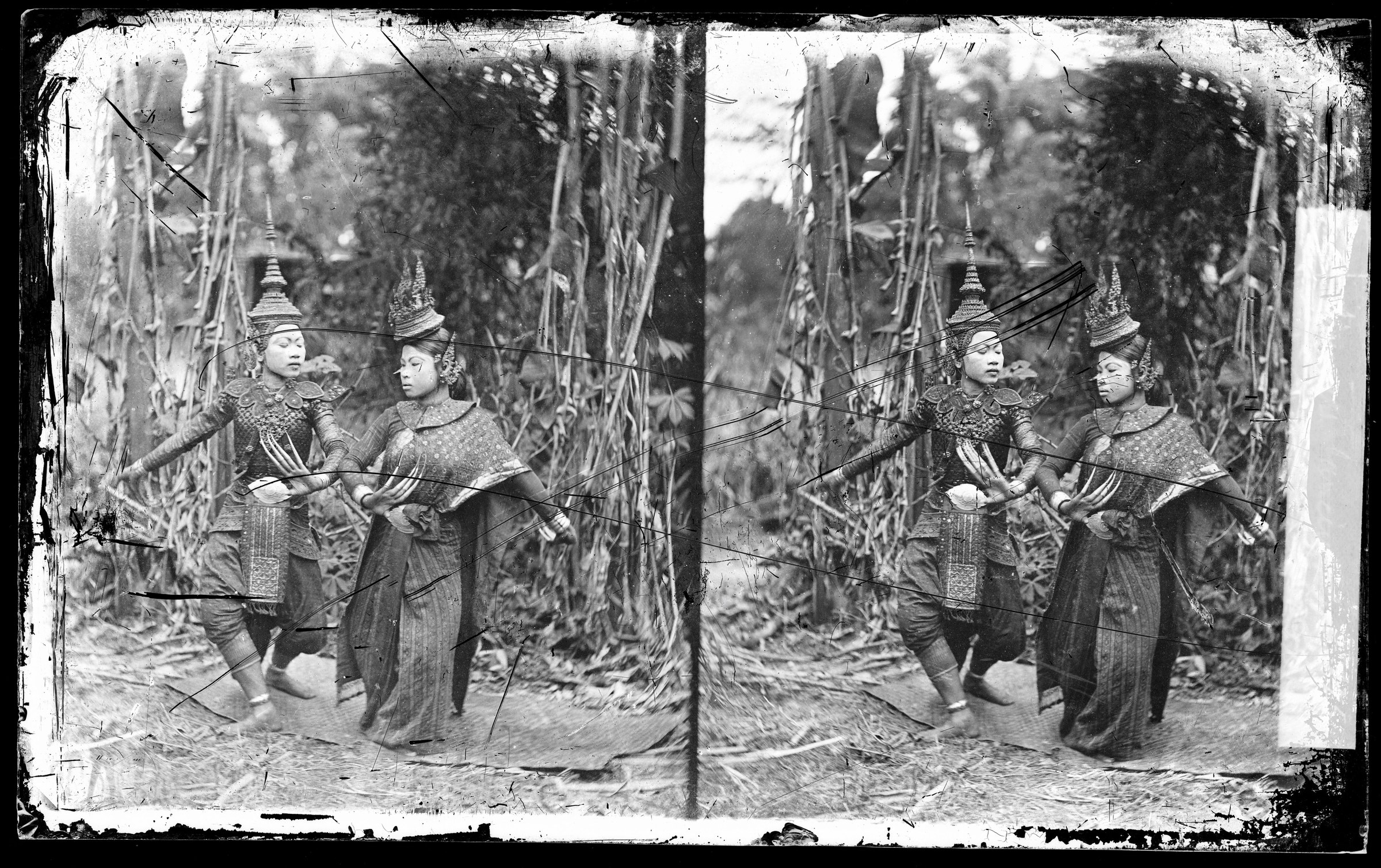 Two Thai dancers in ornate jackets and pointed headdresses pose symmetrically in a forest setting, arms raised in formal gestures.