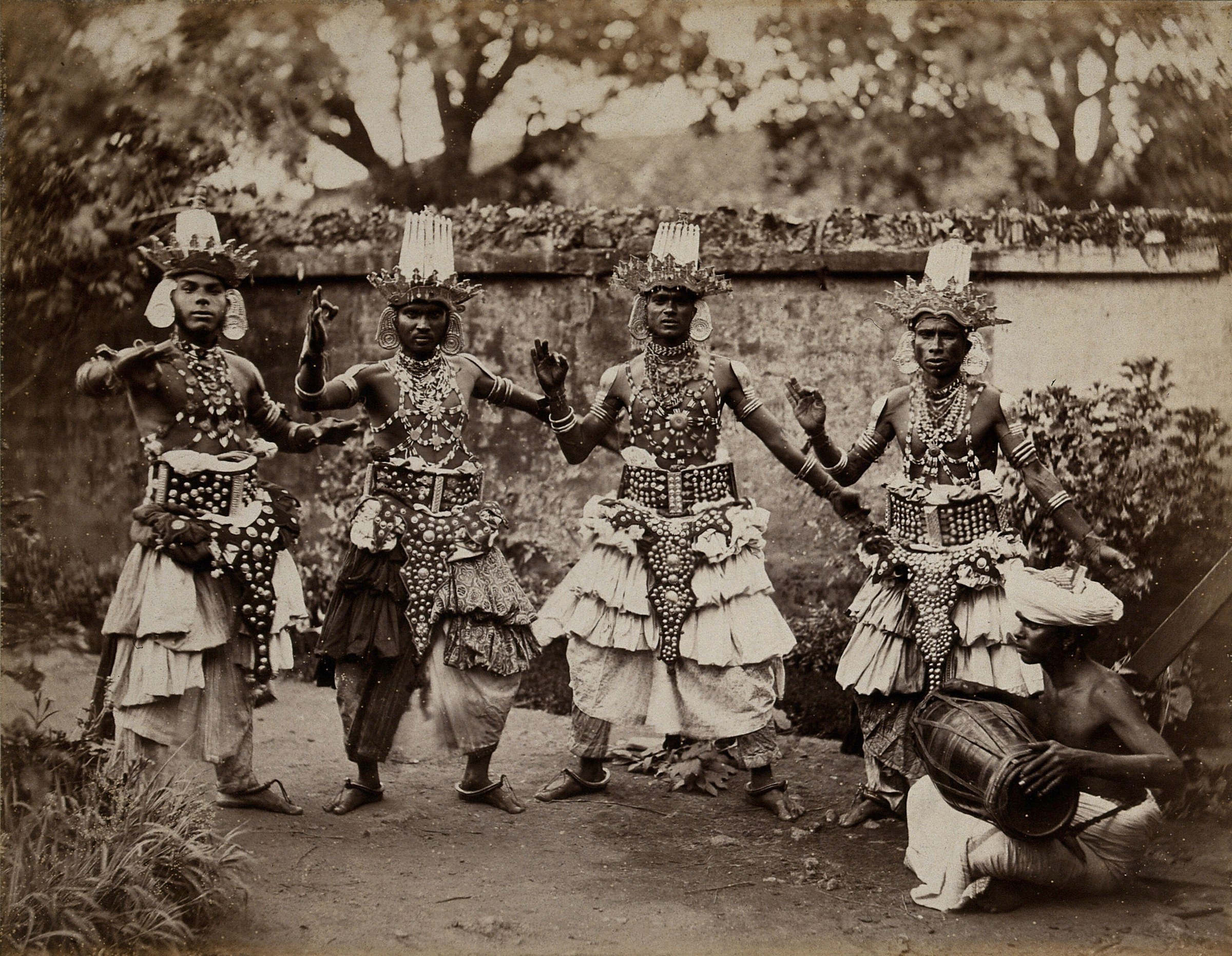 Four elaborately costumed masked dancers stand in a garden with raised arms, wearing tall headdresses and beaded breastplates. A fifth figure holds a drum.