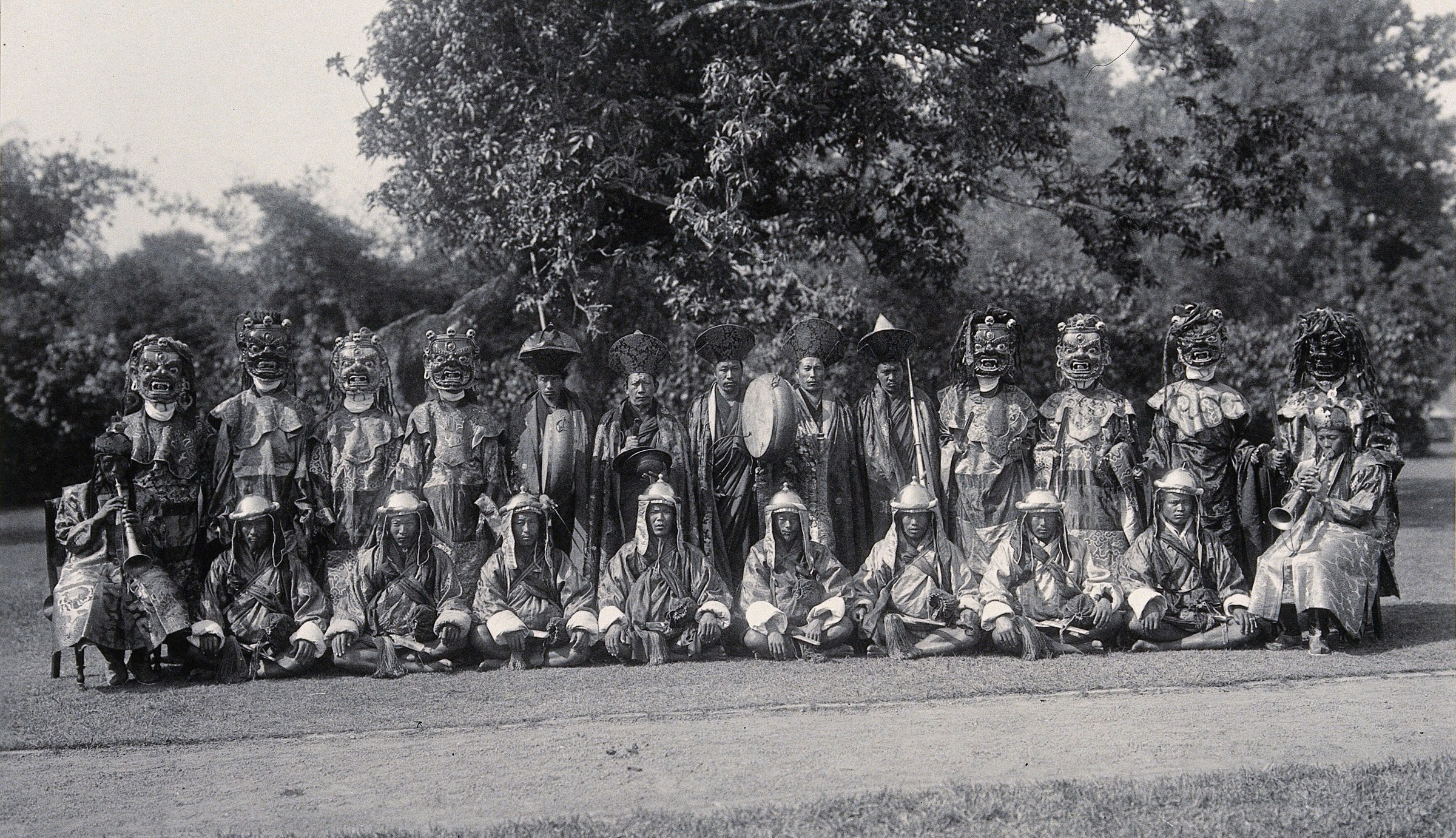 Twenty-five masked figures in elaborate robes and headdresses arranged in two rows on a lawn, facing the camera in formal ceremonial dress.