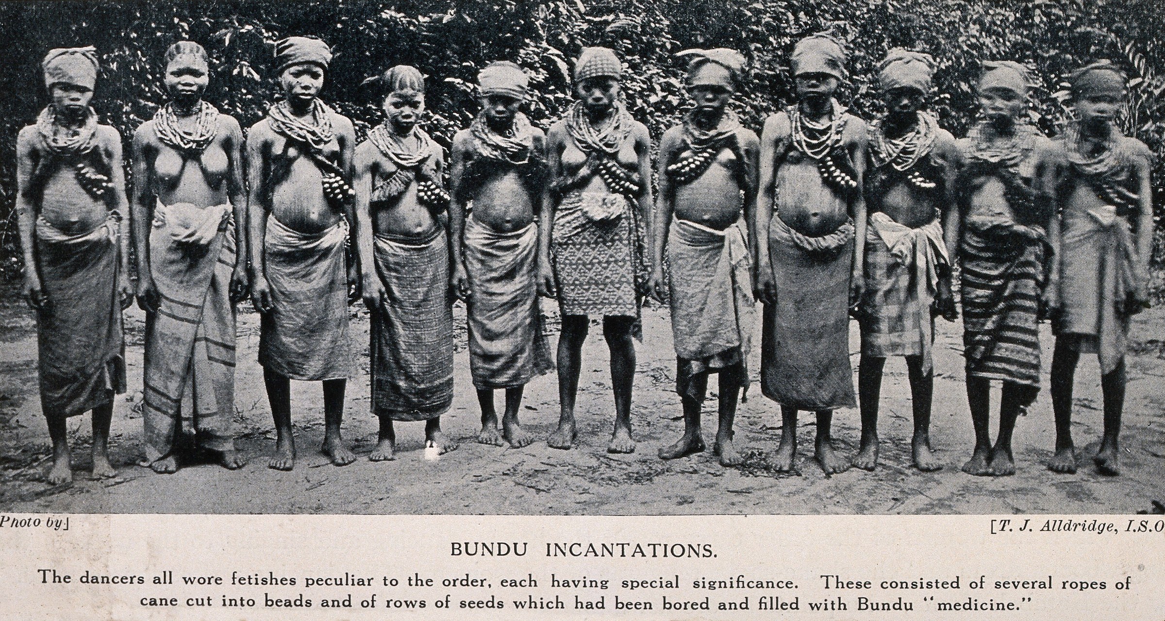 Twelve Bundu women and girls stand in a line wearing beaded necklaces and wrapped skirts, facing the camera in a formal posed group portrait.