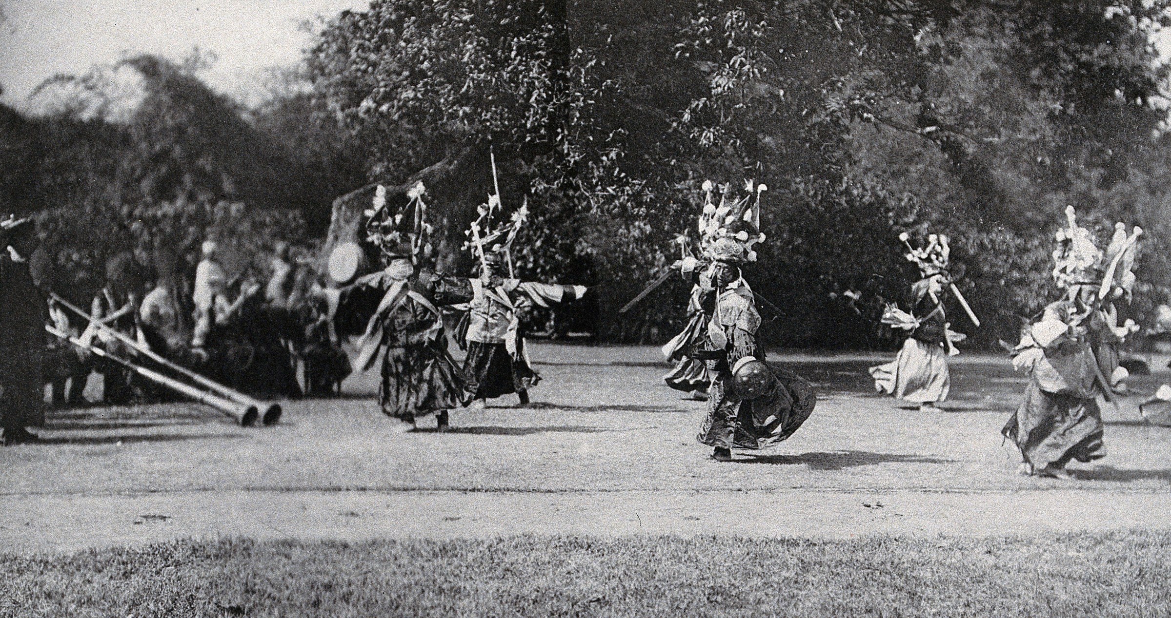 Masked and costumed ritual dancers in elaborate headdresses arranged across a park clearing, photographed in black and white.