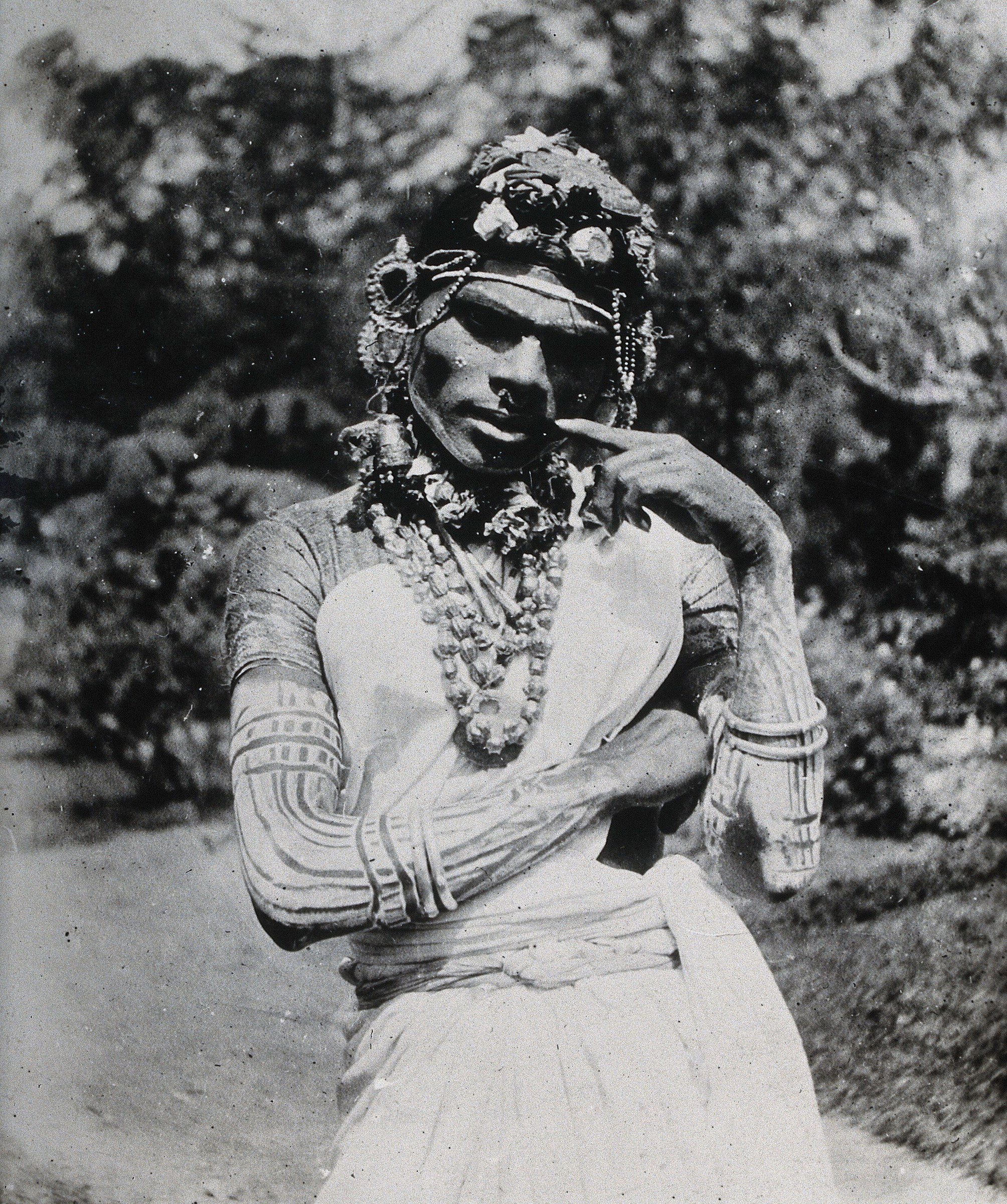 Black-and-white photograph of a Tamil ritual specialist in full ceremonial dress, standing still and gazing upward, adorned with beads, flower garlands, and