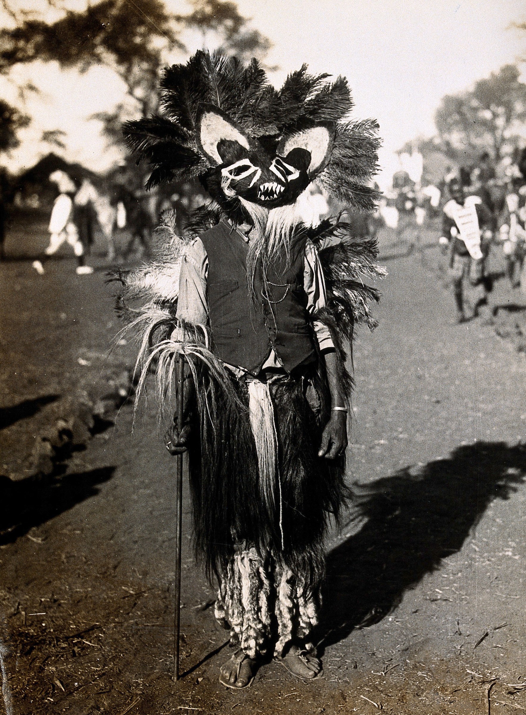 A masked figure in layered black and white costume with feathered headdress stands facing the camera, holding a stick, as onlookers watch in the background.