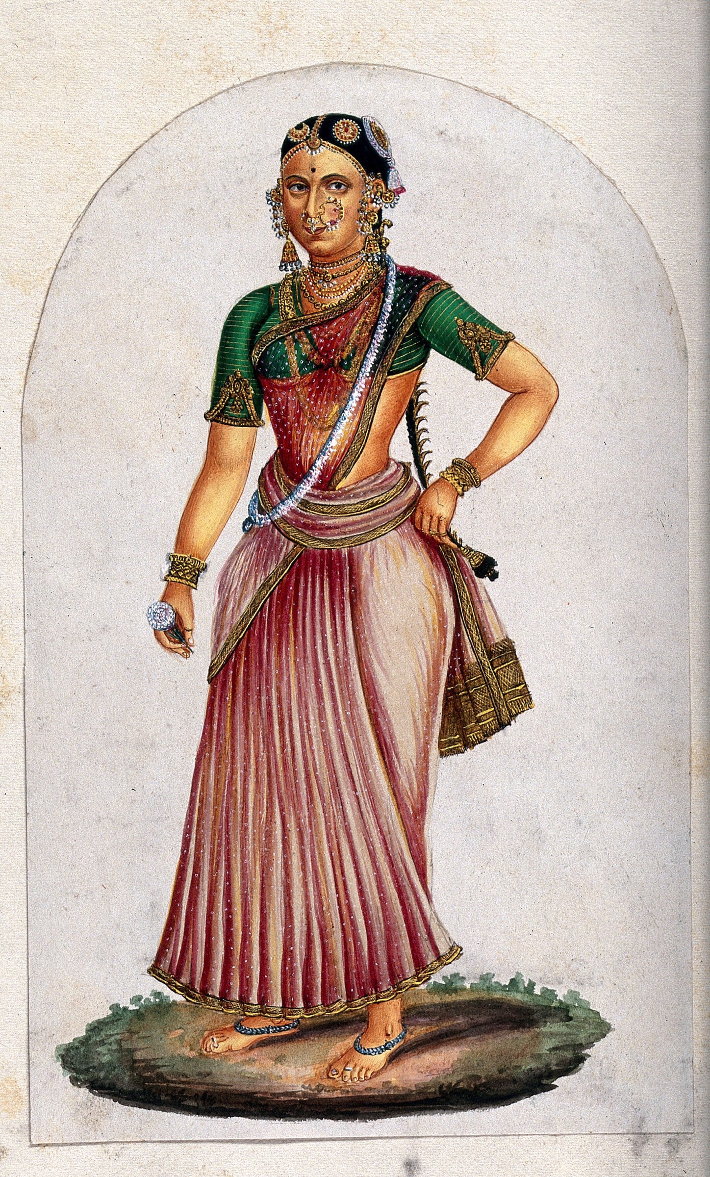 Indian classical dancer in full costume: pleated crimson skirt, green upper garment, extensive gold jewelry, headdress, and facial marking; holding a flower