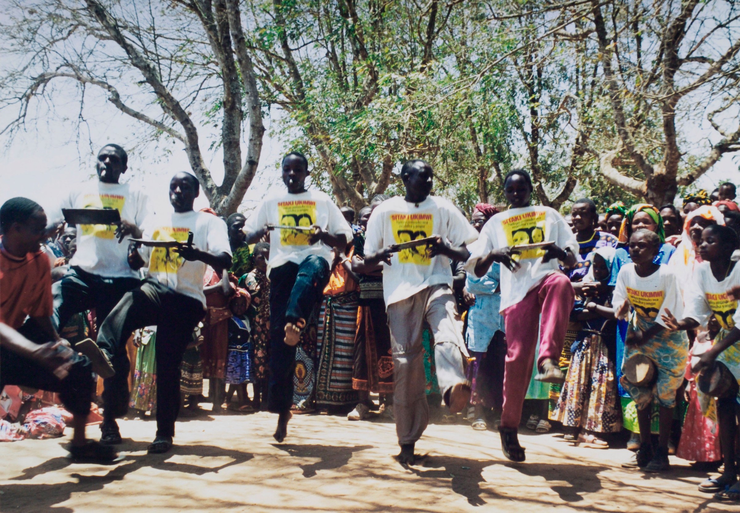 Men in white campaign t-shirts demonstrate movement to a gathered crowd under trees in rural Tanzania.