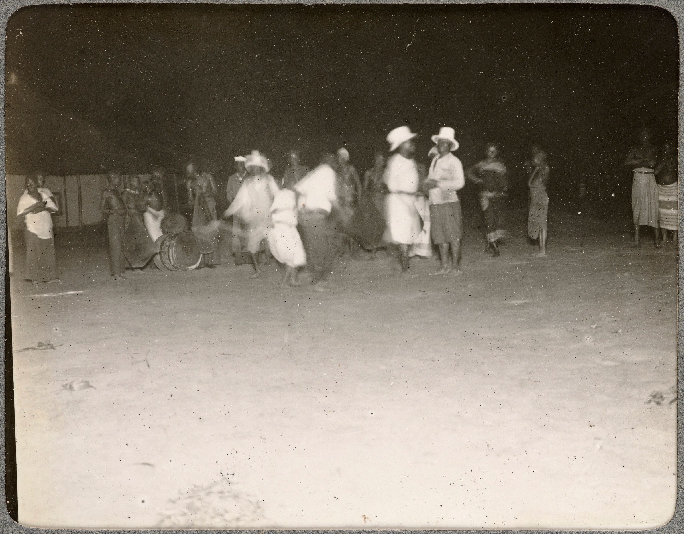 Black-and-white photograph of people dancing outdoors at night; white-clad figures in center with raised arms, watching crowd around them.