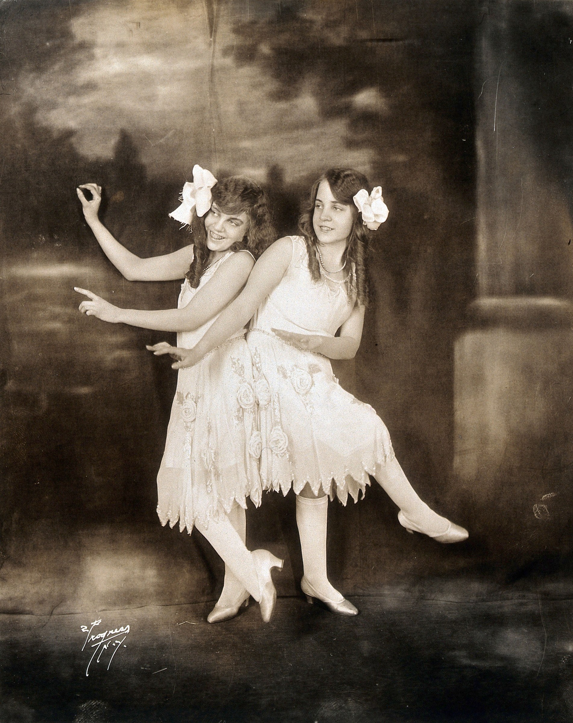 Two young women in sleeveless white embroidered dresses with white bows in their hair, arms extended in expressive gesture, posed in a studio photograph.