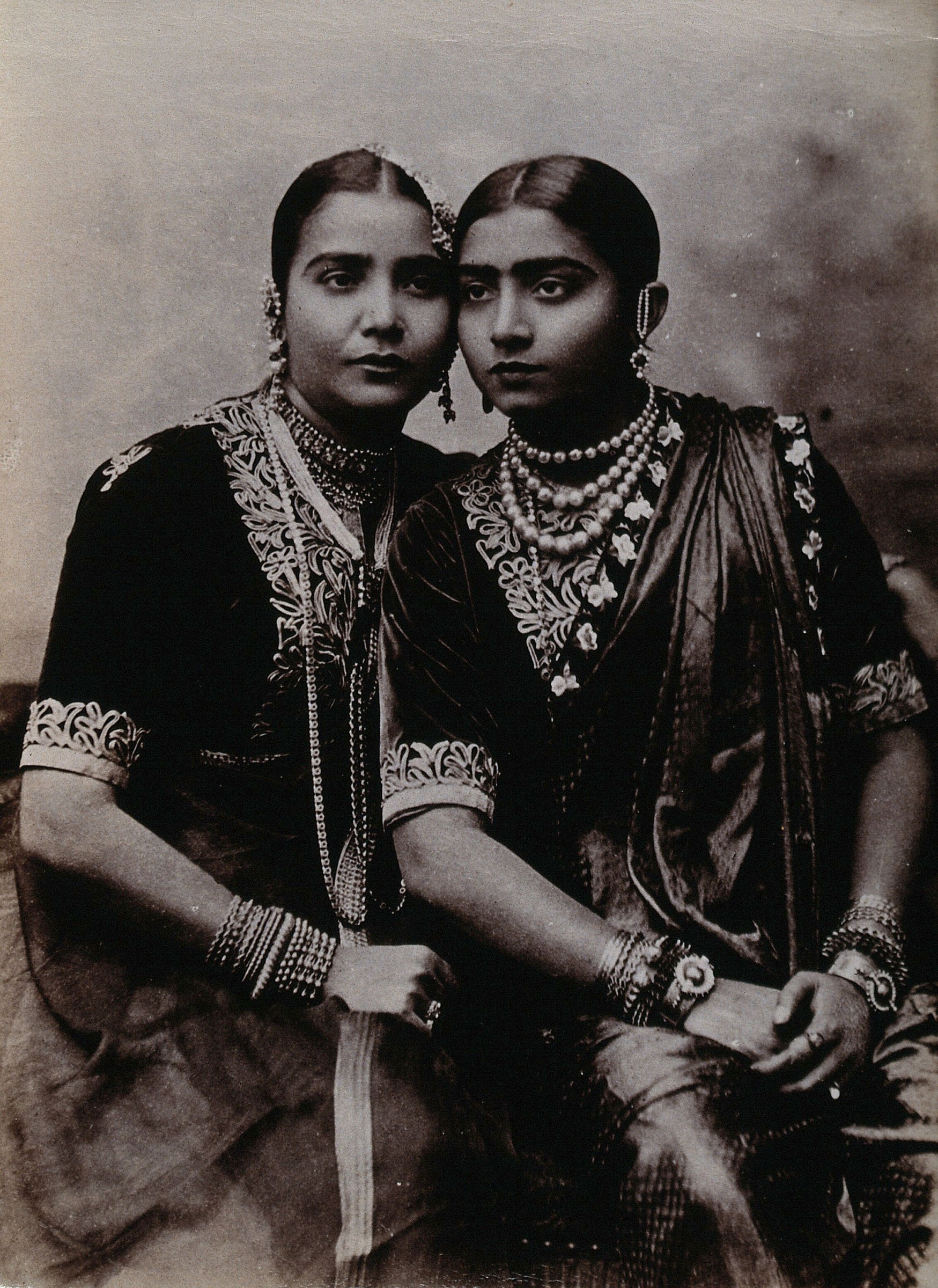 Two women in matching embroidered dark dresses and pearl jewelry stand together in a studio portrait, ca. 1900.