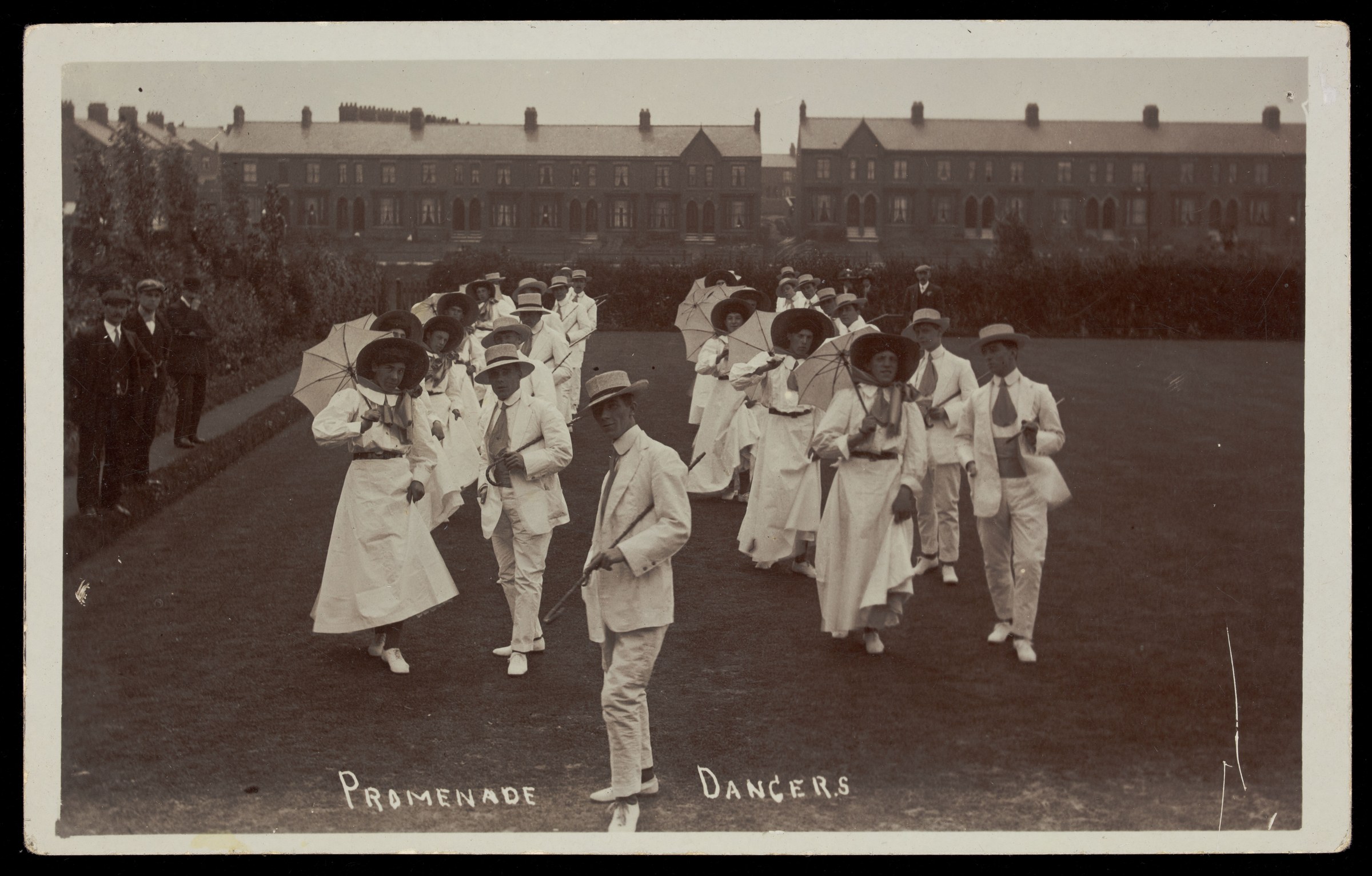 About twenty people in white suits and hats perform a promenade dance outdoors, many holding parasols, with a Victorian building in the background.