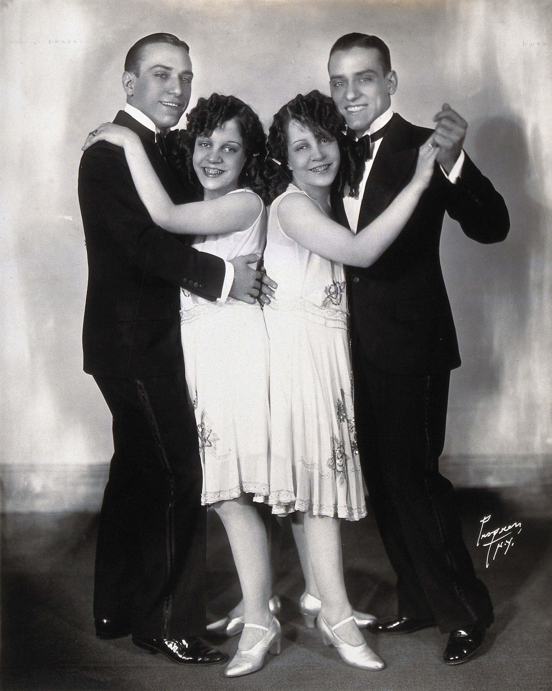 Four performers in formal 1920s evening wear arranged in a symmetrical ballroom dance formation in a studio photograph.
