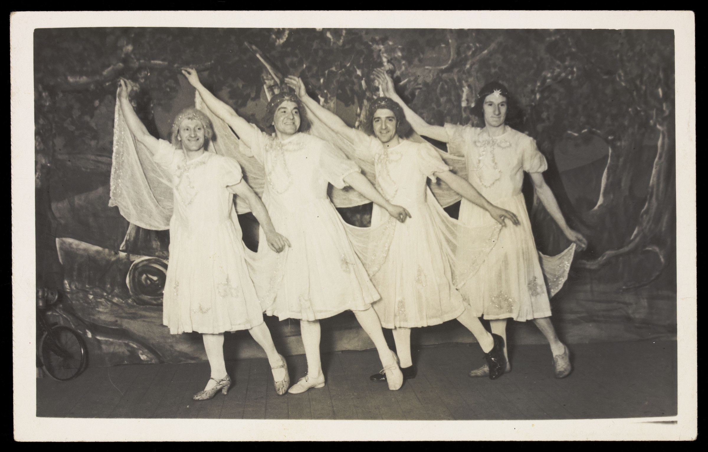 Four men in white dresses stand in a line on a stage, arms raised, in a theatrical pose.