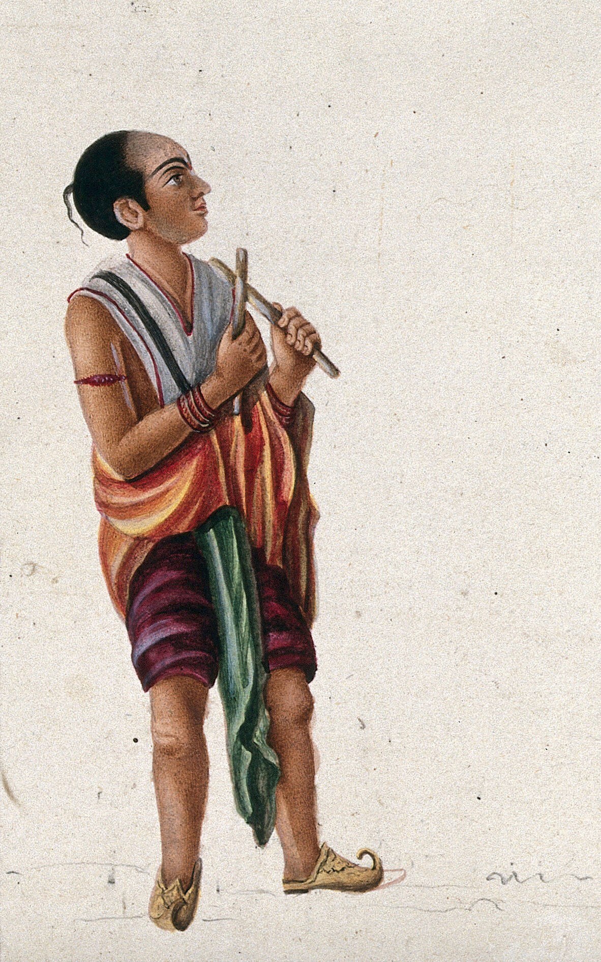 South Asian male dancer in profile, barefoot, holding horizontal wooden sticks (kolu), wearing wrapped skirt and bangles, gazing upward.