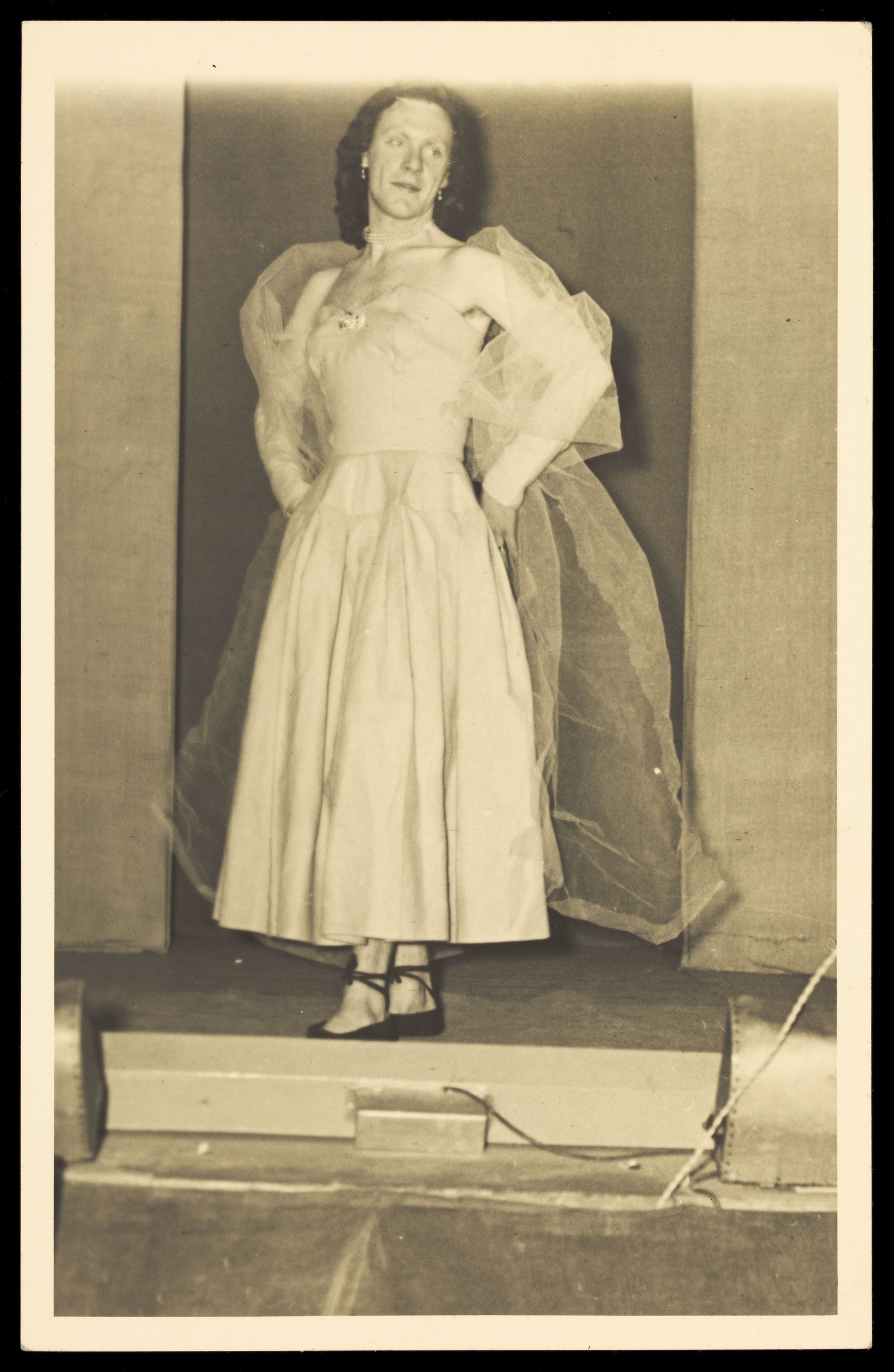 Person in voluminous cream dress and shawl standing upright on a wooden stage, posed frontally for the camera.