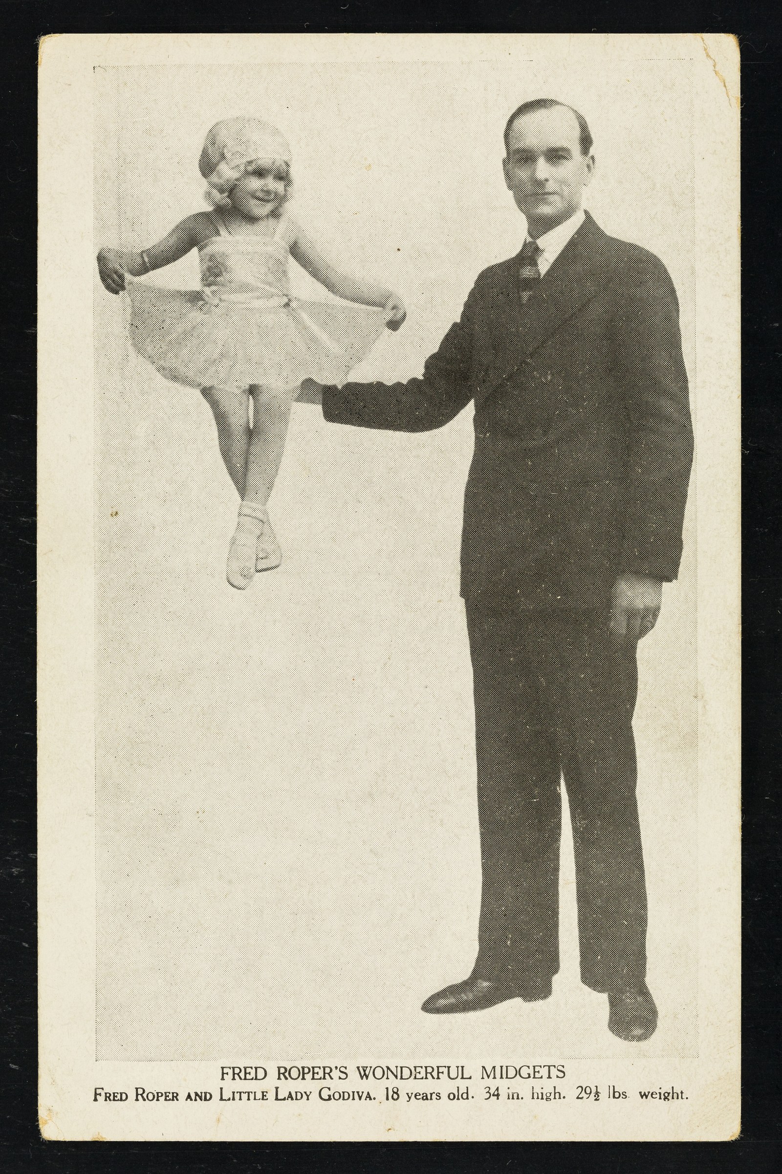 A formally dressed man holds a small child aloft at arm's length. The child extends her arms and legs in a performance pose.