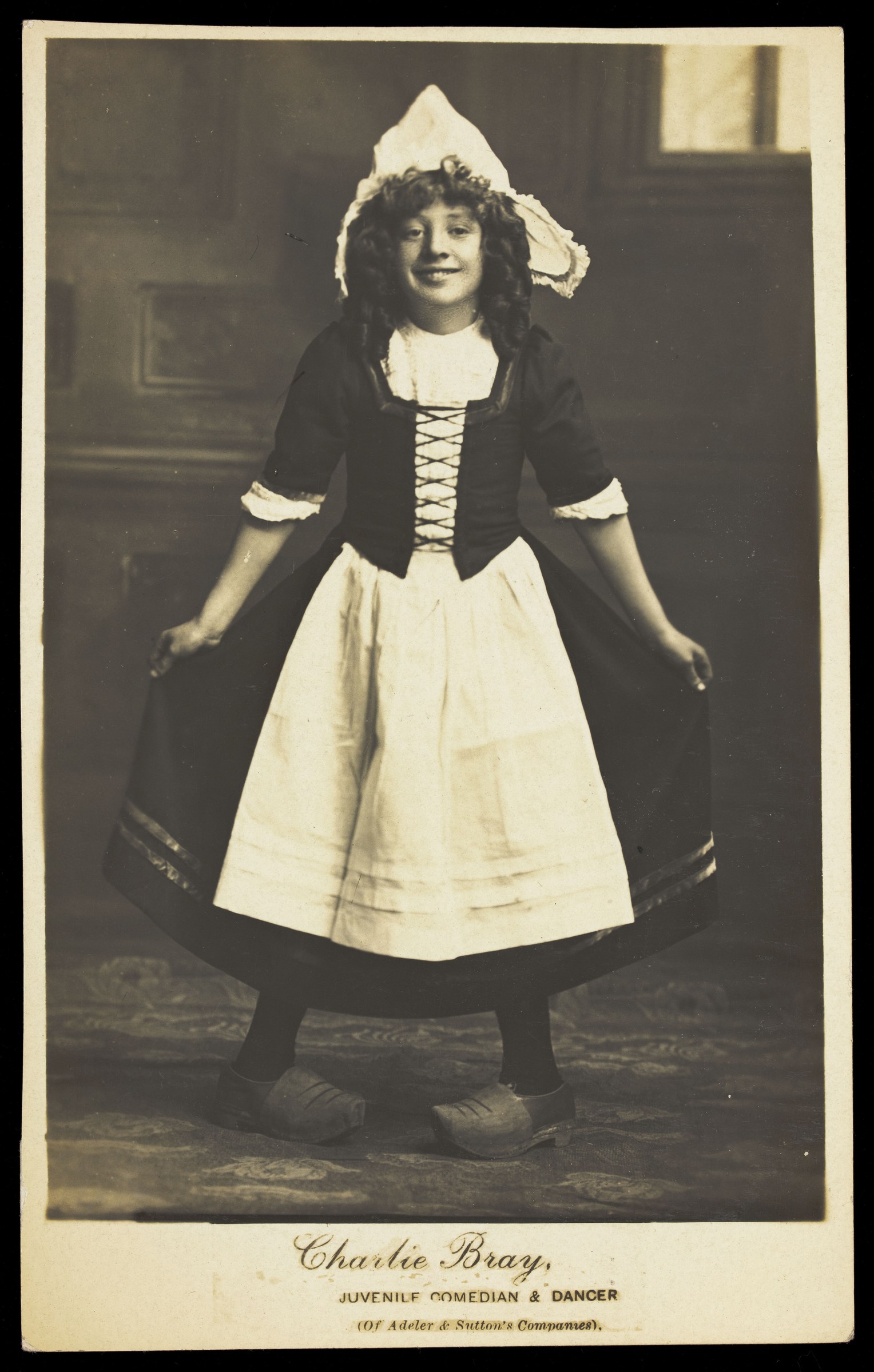 Young performer in Dutch costume—black bodice, white linen skirt, tall white cap, and wooden clogs—standing centered against a dark studio backdrop.
