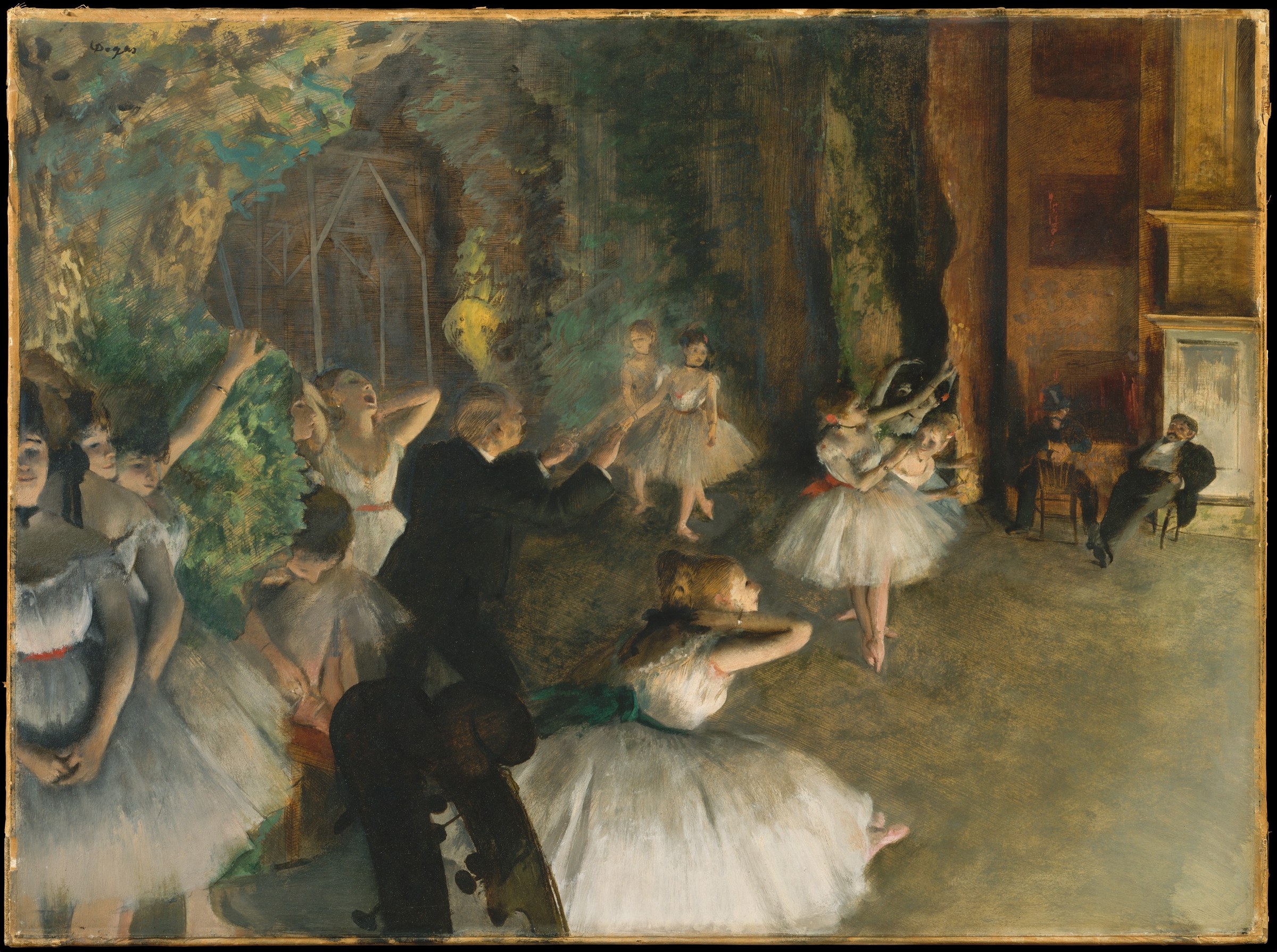 A ballet rehearsal in a pale studio, with a male instructor gesturing toward a group of dancers in white tutus arranged informally around him.