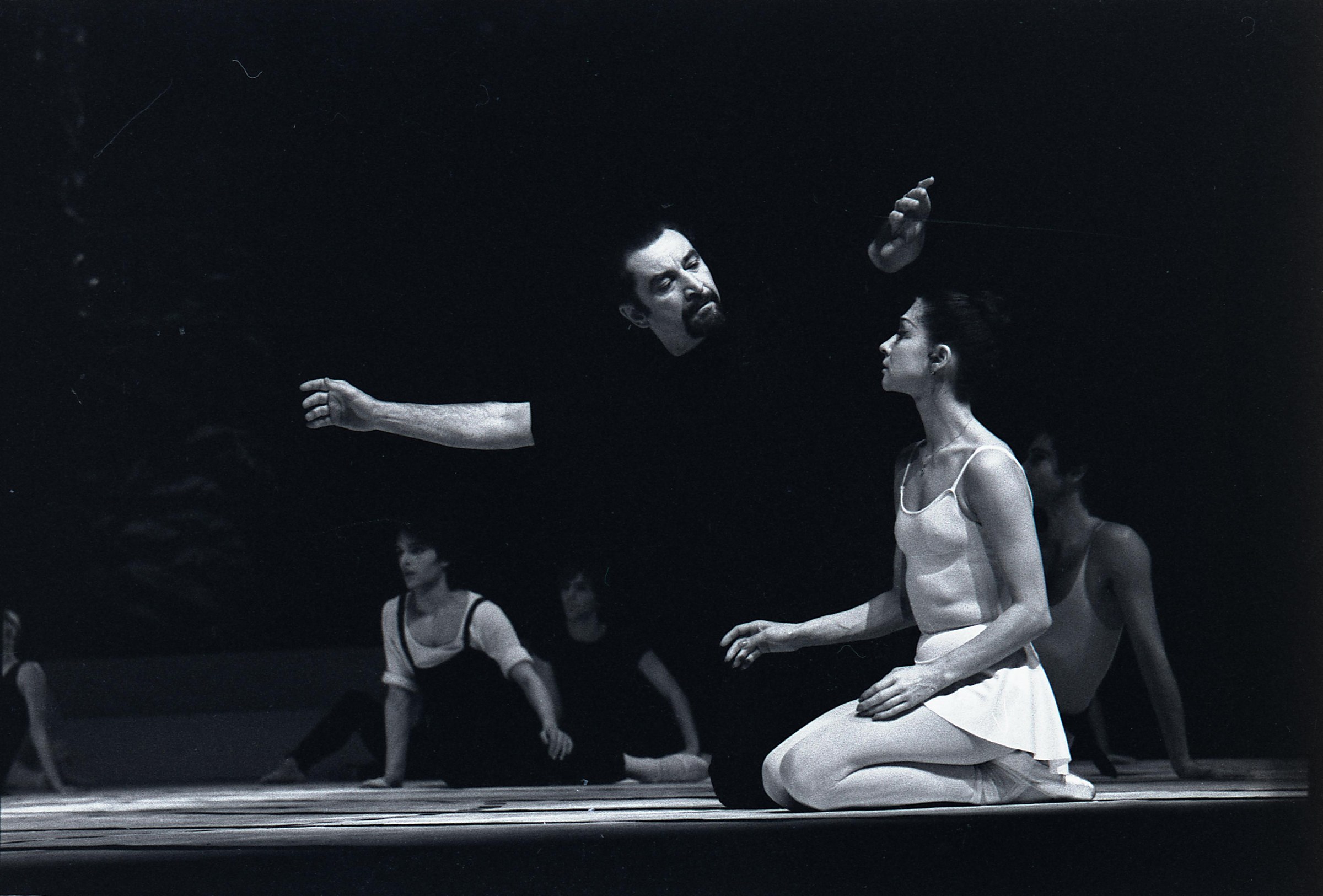 A shirtless male dancer kneels on a dark stage, gazing upward at a second inverted male dancer floating above him, witnessed by seated dancers.