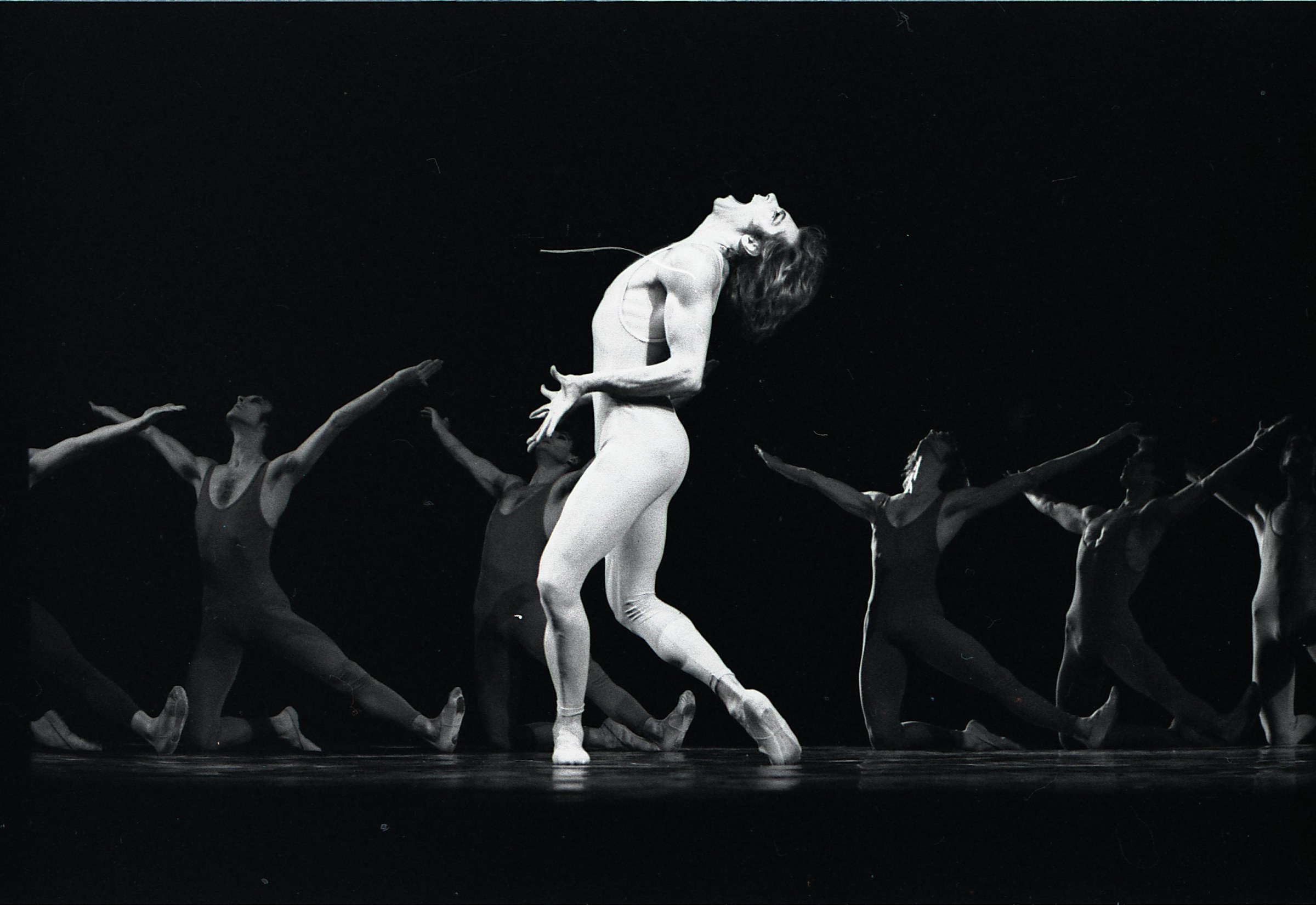 Pale-costumed male dancer in deep backbend, center, surrounded by six dark-clad ensemble members kneeling with raised arms in unison on black stage.