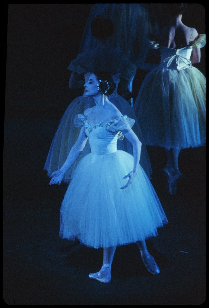 Alicia Alonso in pale tutu and bodice, standing still center-stage in cool blue light, with darker-clad ensemble figures visible in shadow behind her.