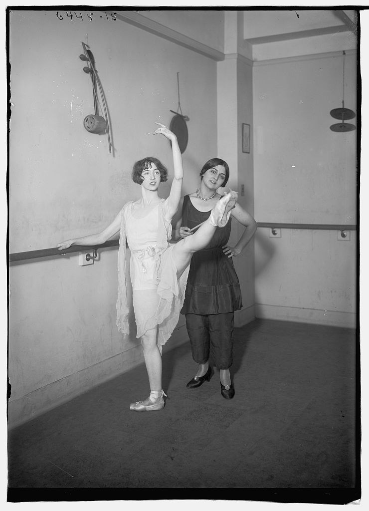 Two women in a bare studio: one performs an arabesque en pointe in a pale tunic with a flowing scarf, while the other stands beside her in a supporting pose.
