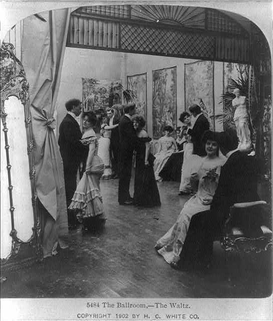 A crowded Edwardian-era ballroom with formally dressed couples; women in white floor-length gowns and men in dark suits among decorated walls and ornamental