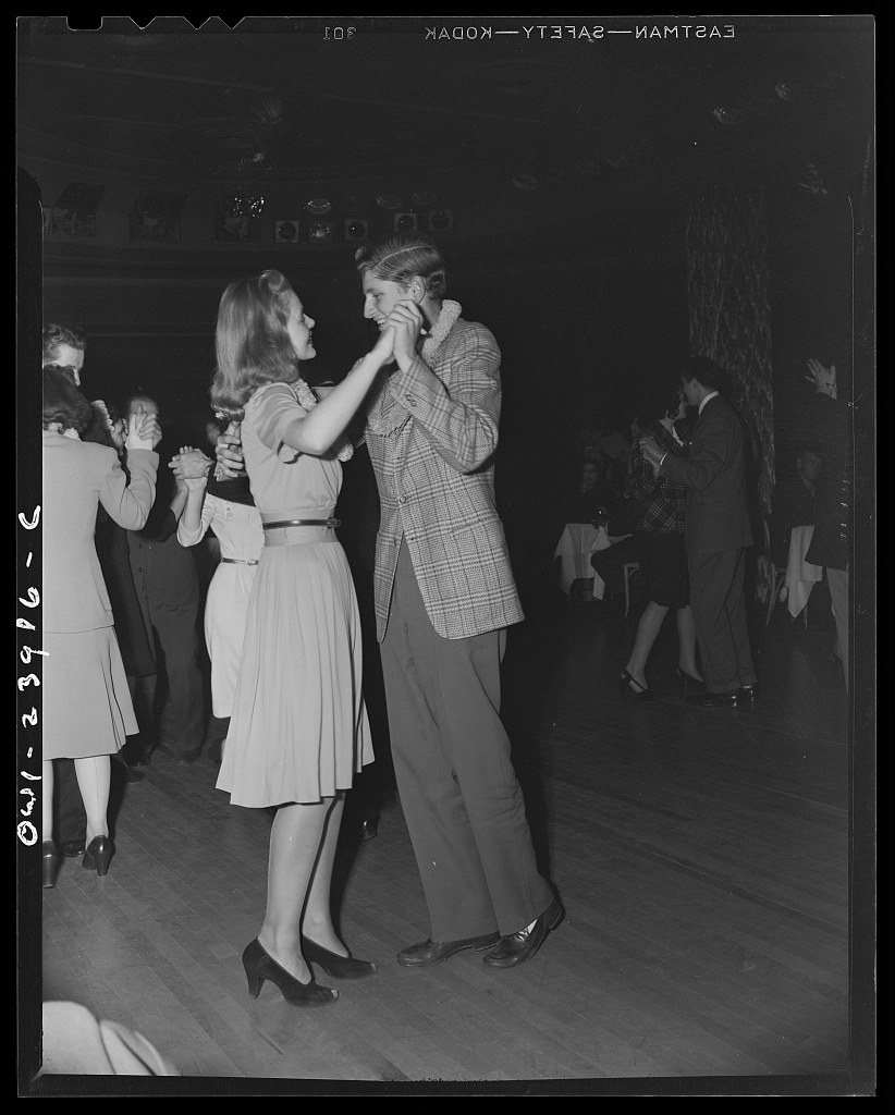 A couple in close embrace dances at a crowded 1940s ballroom, the woman with her head thrown back in joy, surrounded by other dancers.