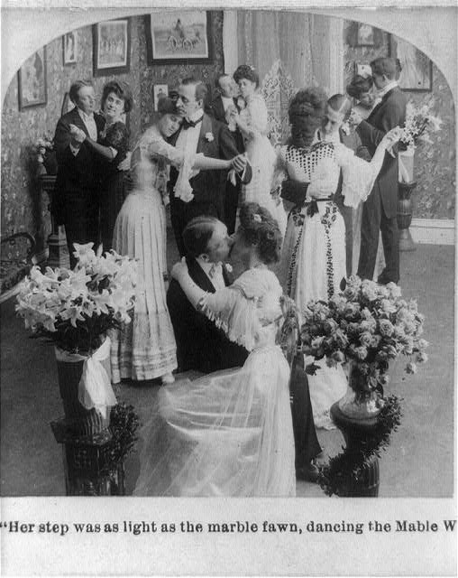 Black and white photograph of a woman in a white gown dancing the waltz with a man in dark suit, observed by seated and standing guests in a flower-adorned
