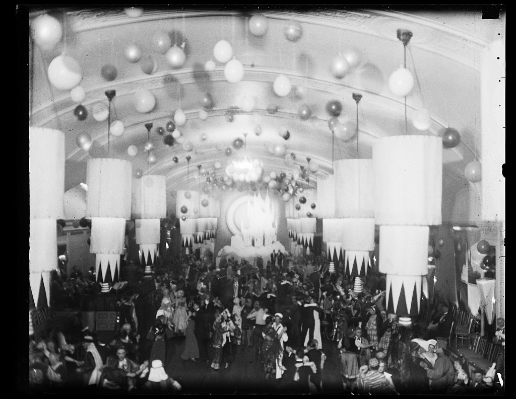 Overhead view of hundreds of formally dressed dancers in a grand ballroom with hanging spheres and tall columns.