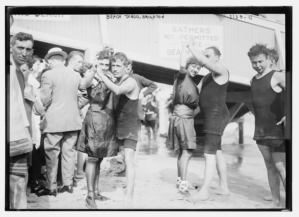 Two couples in 1910s bathing costumes dance tango on Brighton beach, watched by spectators in suits and other swimmers near a beach hut.