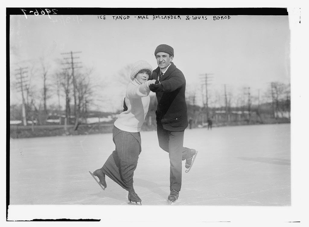 Man in cap lifts woman in white sweater into extended pose on ice, bare trees and utility poles visible in background.