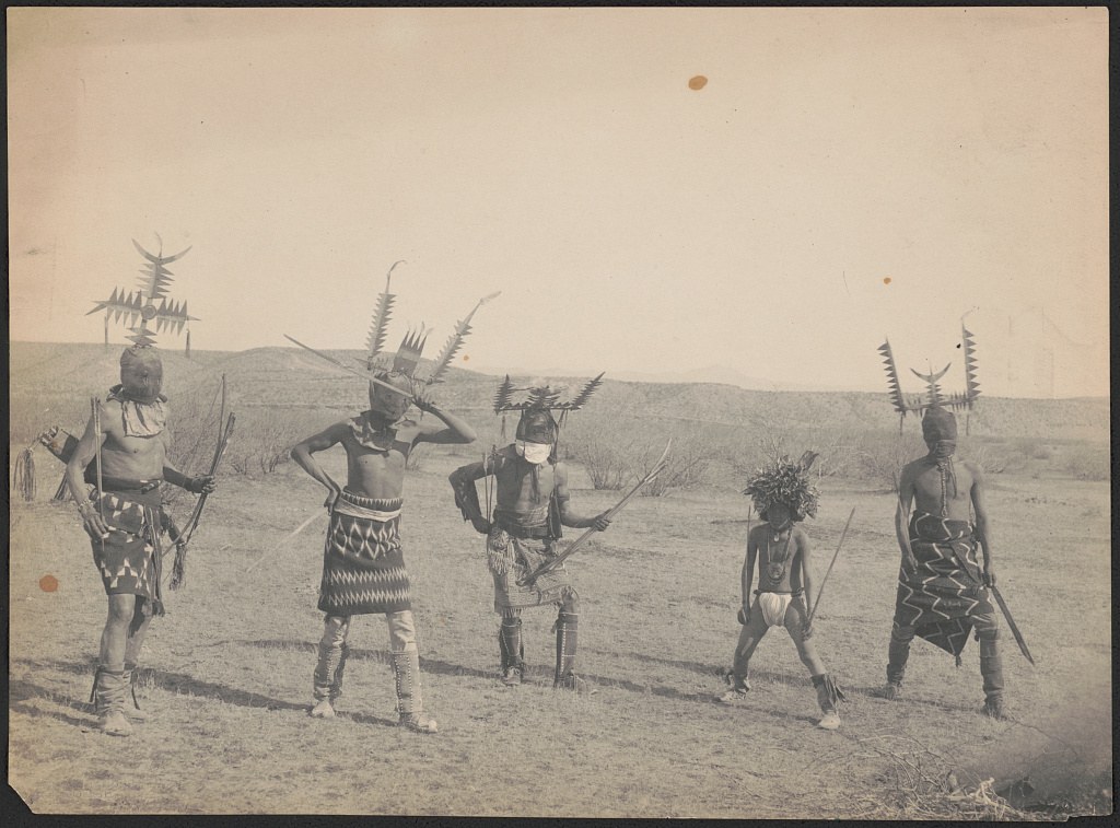 Five men in feathered headdresses and patterned skirts stand in an open landscape holding spears, circa 1899.