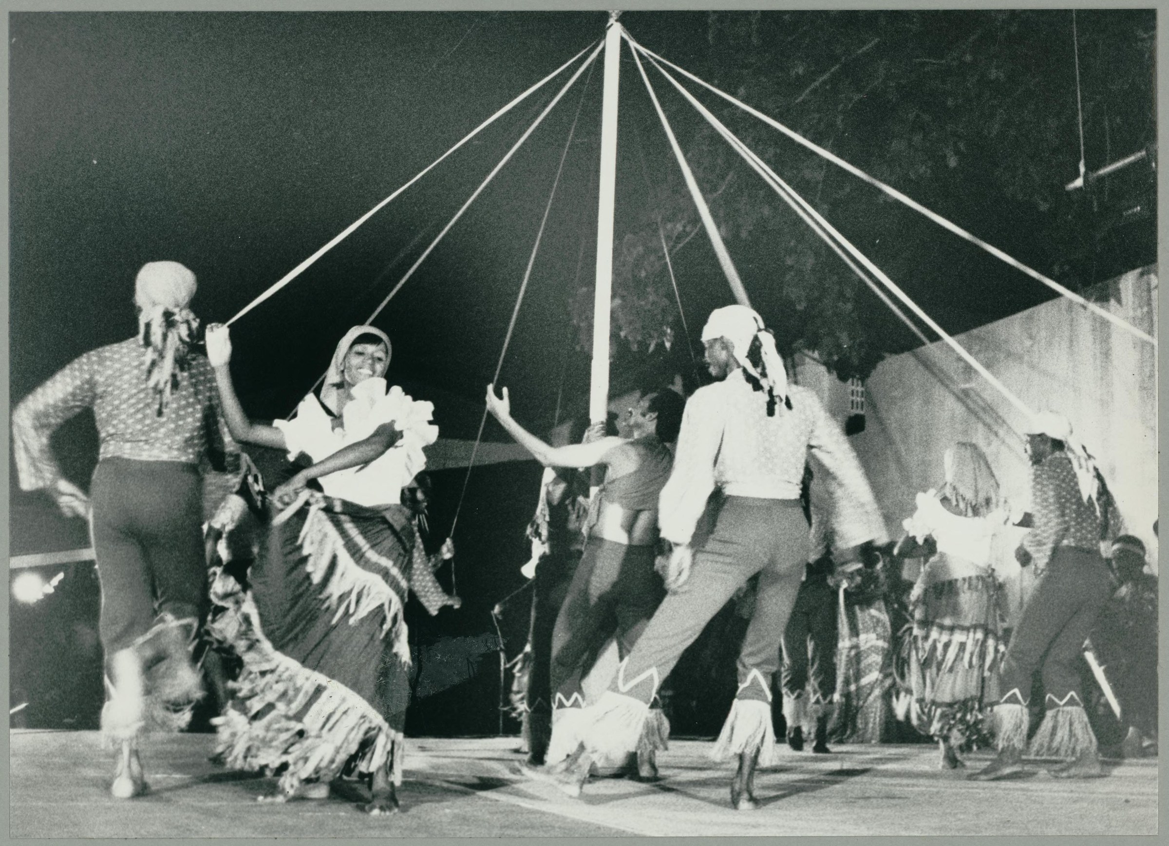 Circle of dancers in patterned costumes holding cords around a central maypole under theatrical lighting, mid-performance.