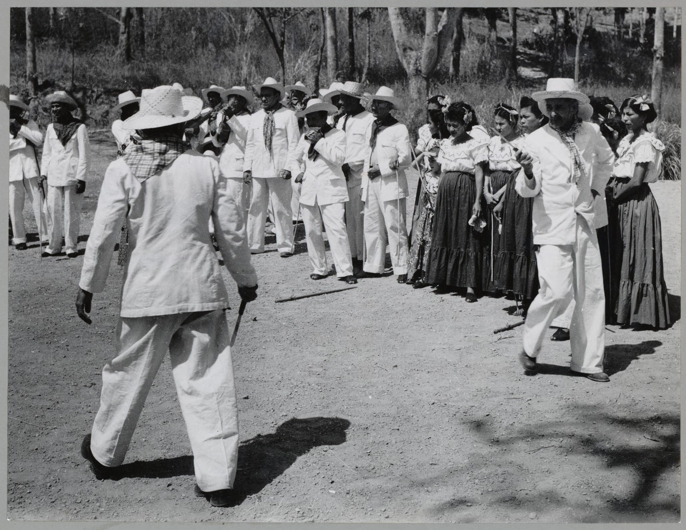 Outdoor daytime gathering of about thirty people in white clothing arranged in a loose circle on a dirt road, with two figures in active motion in the