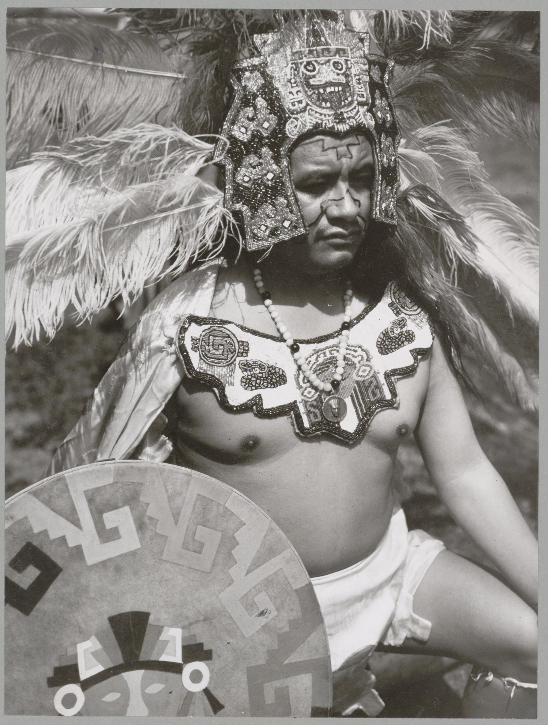 Male dancer in Oaxacan feather dance costume with enormous white feathered wings, beaded headdress, facial paint, bare chest, and decorated shield.