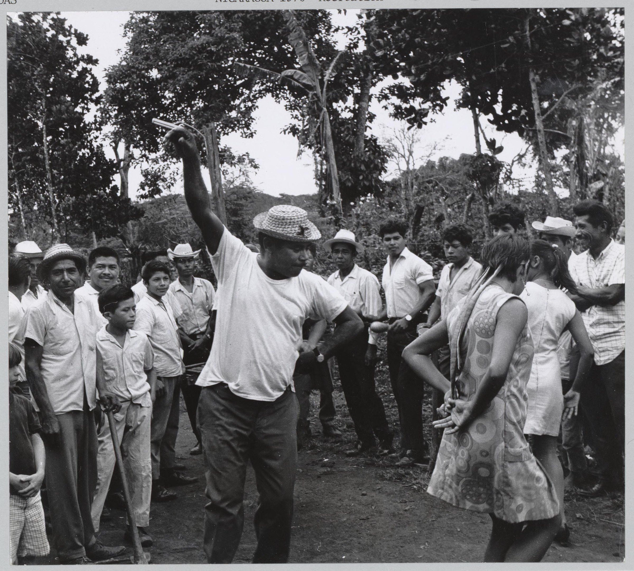 A group of people in white shirts gathered in a forest clearing around a man demonstrating a raised-arm teaching gesture.