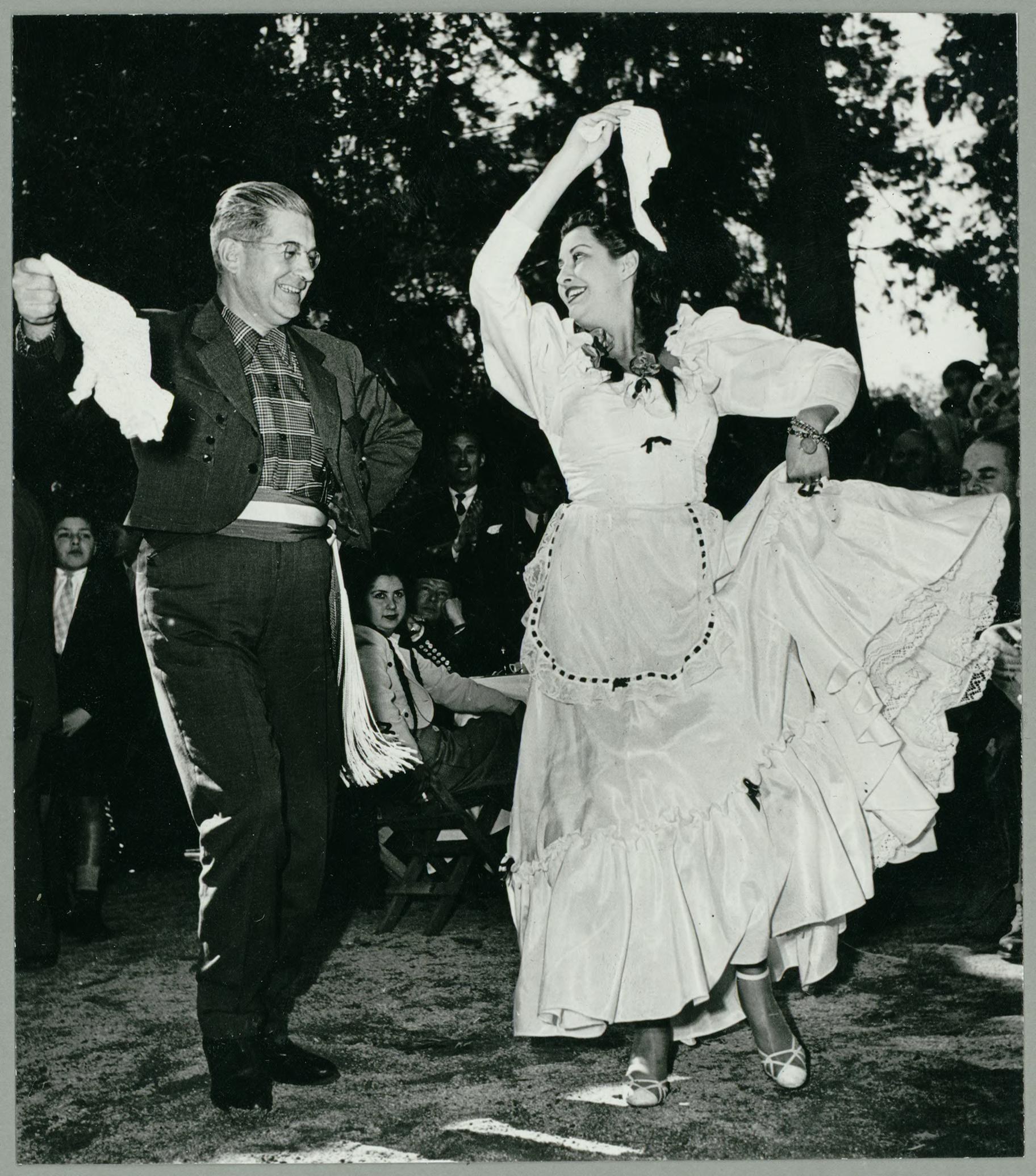 A couple dances outdoors at night under trees, the woman in a white dress and the man in a dark suit, both holding white handkerchiefs aloft.