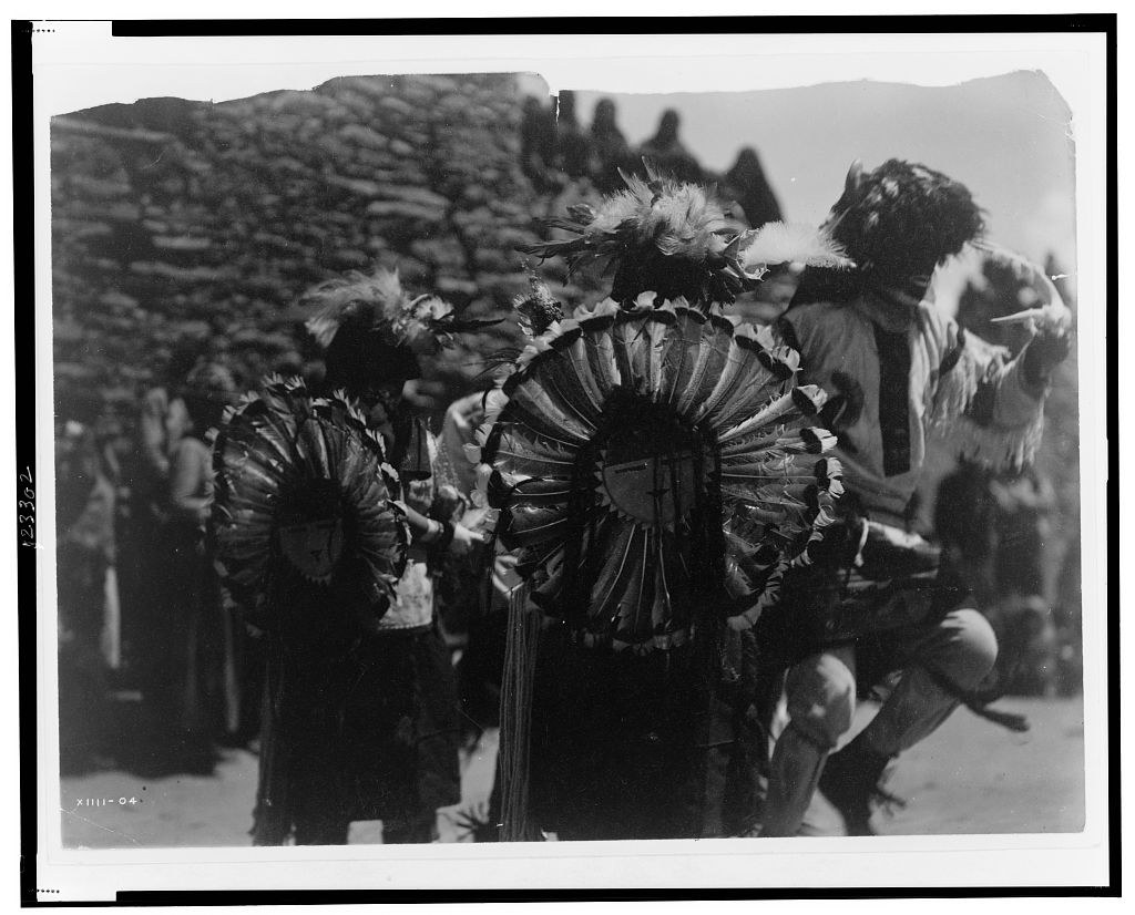 Three Native American dancers in feathered bustles and fringed regalia stand outdoors with forested mountains in the background, posed for the camera.