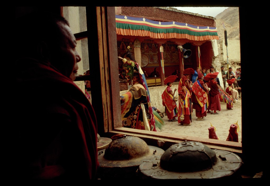 Buddhist monks in red robes stand in formation in a mountain monastery courtyard, facing a decorated ceremonial platform with religious iconography.