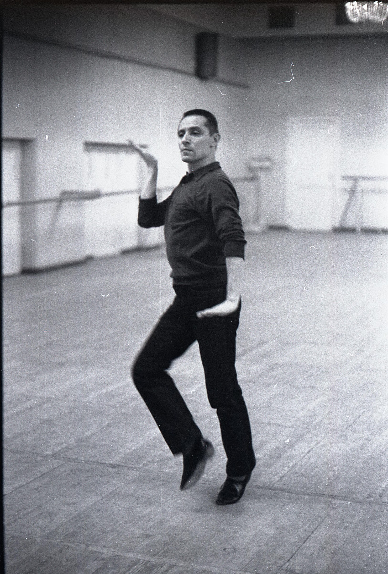 Male ballet dancer suspended in mid-air, body spiraling, one leg extended upward, arms reaching, in black studio wear against white wall.