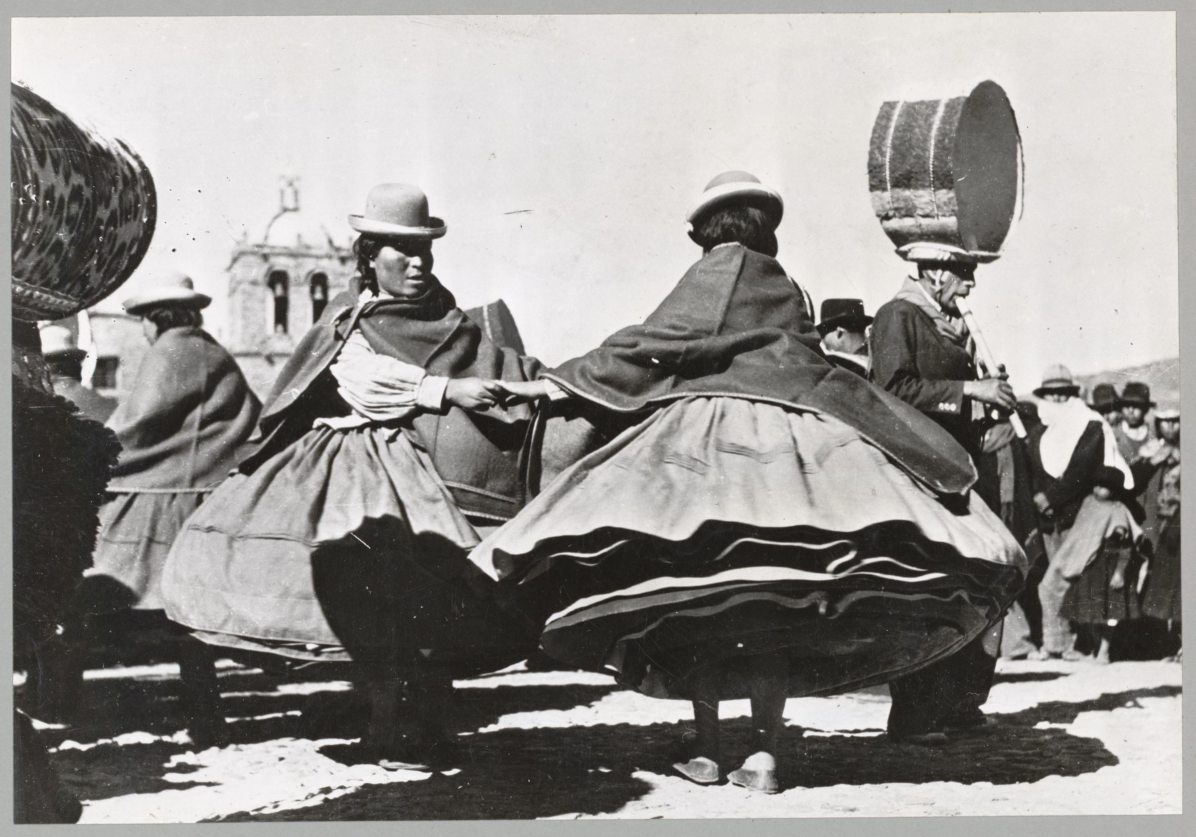 Two women in layered skirts and bowler hats dance with large cooking pots in an Andean plaza before a colonial church, 1960.