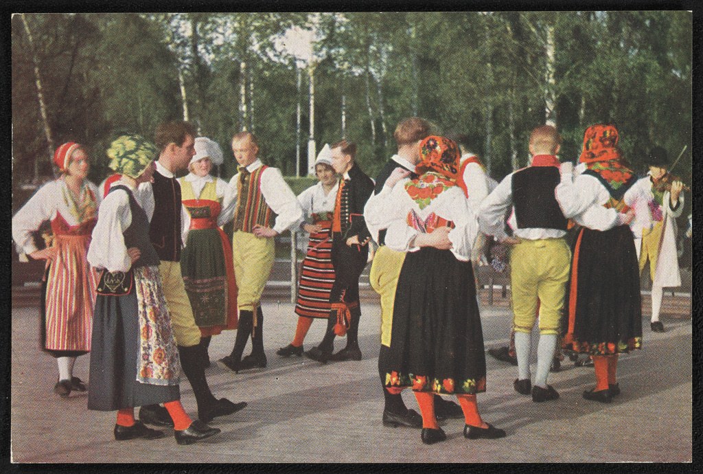 A group of fifteen people in colorful Scandinavian folk costumes stand outdoors among birch trees at Skansen museum in Stockholm, circa 1900.