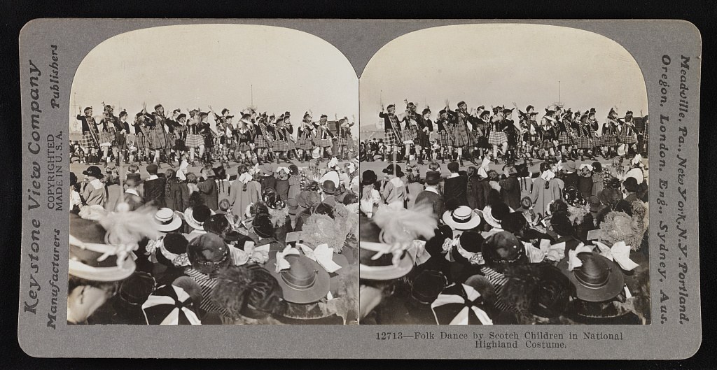 Stereoscopic photograph of Scottish children in Highland dress standing on a platform with crowds of spectators seated and standing below them outdoors.