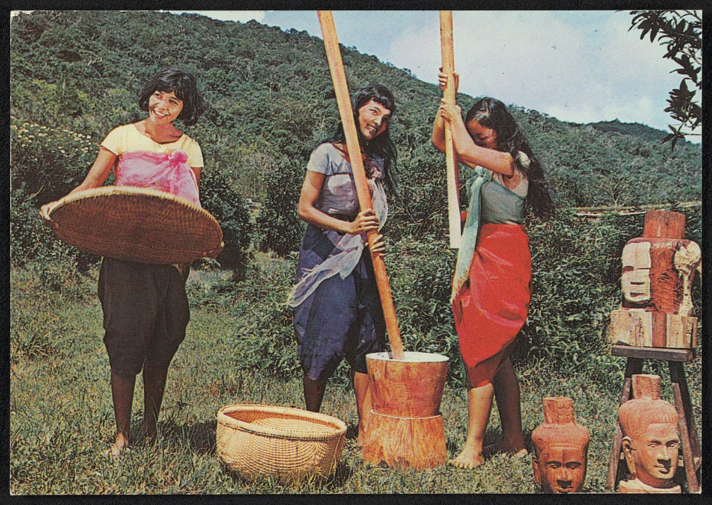 Three bare-footed women in sarongs process grain outdoors using woven baskets and wooden poles, with carved wooden figures visible to the right.
