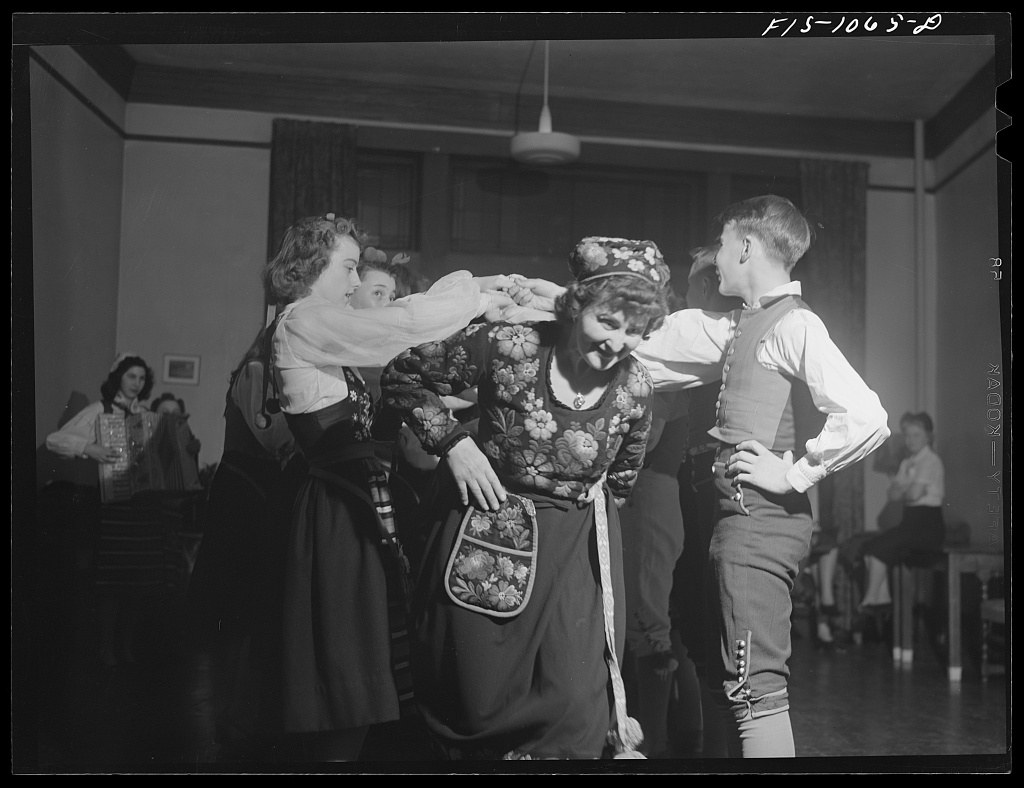 An older woman in traditional floral dress teaches folk dance to four children in a community hall, 1942.